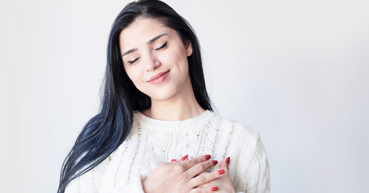 Frau hält mit meinen Händen ihre Herzgegen fest und lächelt. Sie praktiziert Selbstfürsorge